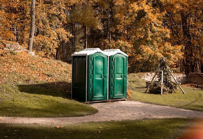 Two green portable toilets stand in a park with a backdrop of trees filled with orange leaves.